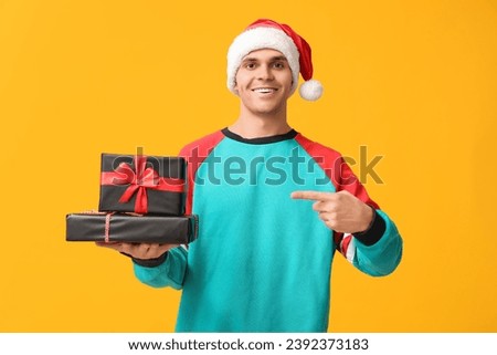 Handsome young man in Santa hat pointing at Christmas gifts on yellow background