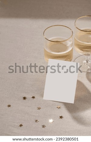 Holiday greeting postcard or invitation template, blank paper card mockup, wineglass with sparkling wine on neutral beige linen tablecloth background with gold confetti and natural shadows, lifestyle.