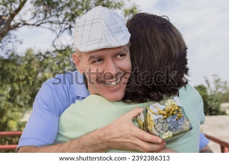 Grey haired dad and adult son. Fathers Day. Parent is holding present box and hugging Young man with long hair outdoors in sunny day. Concept of family, relationship, holiday. Royalty-Free Stock Photo #2392386313