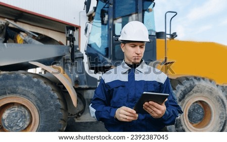 Industrial worker young man in helmet holds tablet computer on background of production of excavator factory.