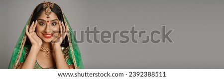 joyful indian woman with bindi dot and green veil smiling at camera with hands near face, banner