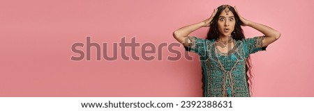 amused young indian woman in traditional costume with hands on head posing on pink backdrop, banner