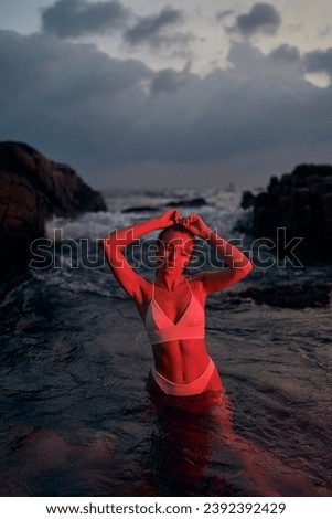 Attractive woman enjoys nocturnal swim in ocean, illuminated by red light. Fashionable female in swimwear relaxing in sea at evening. Nighttime beauty, leisure by beach silhouette. Stylish lady bathes