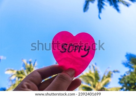 The word sorry written on a pink sticky note in the shape of a heart isolated on a blue sky background. Concept of Apologizing with a sky background