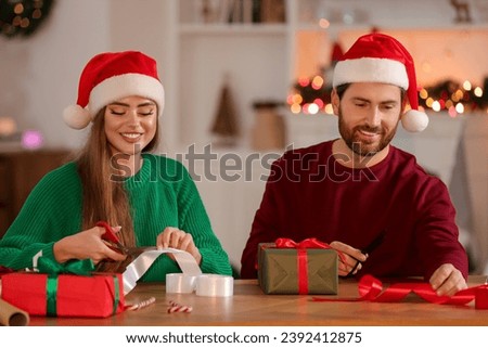 Happy couple in Santa hats decorating Christmas gifts at table in room