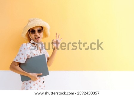 half body photo Asian business woman wearing glasses, hat and dress, holding a laptop, making OK sign with hand and saying wow, relaxing at vacation hotel near yellow wall in beautiful location.