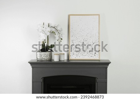 Picture frame, candles and orchid on fireplace near white wall indoors. Interior design