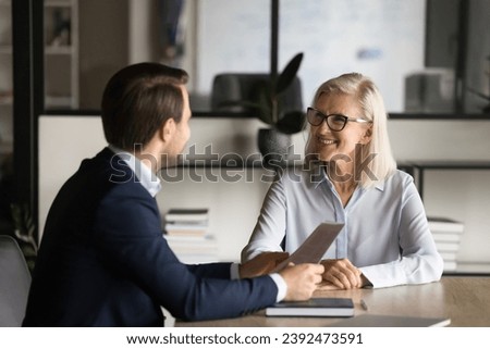 Two happy older and younger business partners speaking at meeting table, discussing paper contract. Employer and candidate talking on job interview, negotiating on resume Royalty-Free Stock Photo #2392473591