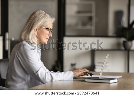 Serious blonde elder business woman in elegant glasses typing text on laptop, sitting at workplace table, using computer for online work communication, wireless Internet connection