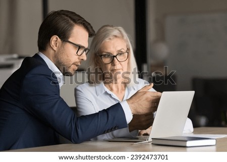 Two serious business colleagues collaborating on Internet project at laptop, watching online content at computer, pointing hand at screen, speaking. Software expert explaining client application work Royalty-Free Stock Photo #2392473701
