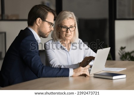 Focused senior business professional complaining on laptop, software work problem to application developer, computer expert, sitting at laptop, pointing at display, speaking Royalty-Free Stock Photo #2392473721