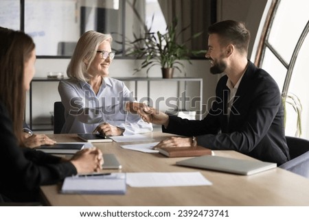 Positive successful elder businesswoman shaking hand to younger best worker man on corporate meeting. Happy diverse managers working on business collaboration, teamwork, partnership