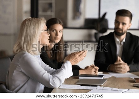 Confident blonde senior business professional woman talking to younger colleagues on brainstorming meeting, offering ideas for work on project, discussing strategy, management