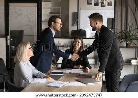 Two happy confident business partners men shaking hands over large table, meeting with project colleagues, managers, closing deal, contract, discussing investment to startup, partnership