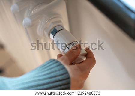 Hand adjusting heater knob, regulating temperature at home in winter during heating season. Radiator equipped with thermostat allowing to save, economize energy, limit temperature in apartment Royalty-Free Stock Photo #2392480373