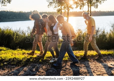This lively image depicts a group of friends playfully walking along a lakeside path as the sun sets. Their movement conveys a sense of energy and carefree enjoyment. The warm sunlight flares through Royalty-Free Stock Photo #2392488927