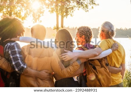 The image captures a poignant moment of four friends, seen from behind, embracing as they look out over a lake bathed in the soft light of the setting sun. The sun's rays filter through the trees Royalty-Free Stock Photo #2392488931