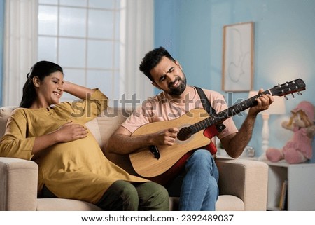 relaxed indian pregnant woman listening to husband song by playing guitar while sitting on sofa at home - concept of Maternal comfort, relationship bonding and entertainment