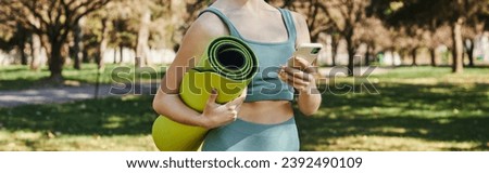 cropped banner of sportswoman in active wear holding smartphone and fitness mat in green park