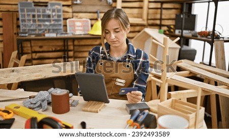 Attractive young blonde female carpenter, engrossed in serious business of financing her carpentry workshop, using credit card  touchpad amidst timber, indoor