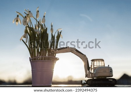 bouquet of snowdrops in a small bucket and a toy excavator in backlight against the backdrop of the sunset sky. Builder's Day holiday, construction business card