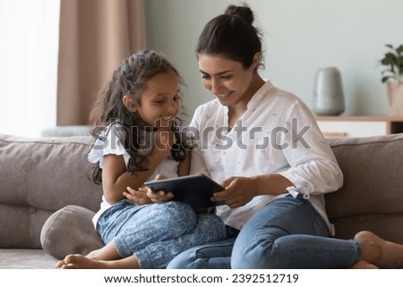 Young Indian mom show little daughter new cool application on digital tablet for development, family spend leisure at home with modern wireless device, enjoy fun on-line. Generation z use tech concept
