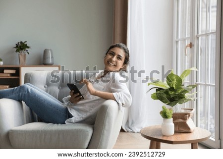 Cute Indian woman relax on armchair smile use smartphone. Buyer of on-line goods, social media or new mobile application usage for comfort life. Electronic commerce services user, modern tech concept Royalty-Free Stock Photo #2392512729