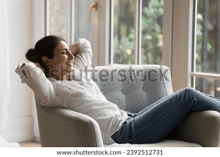 Calm Indian woman closed eyes spend carefree weekend, enjoy lazy leisure, breath fresh conditioned air inside cozy room. No stress, climate control, smart home, modern furniture, comfort life concept Royalty-Free Stock Photo #2392512731