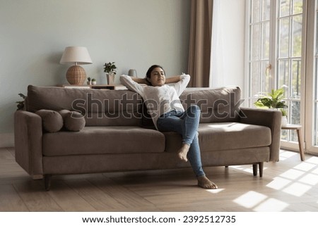 Inside clean cozy light living room young Indian woman resting on sofa put hands behind head closed eyes enjoy day off and fresh conditioned air at modern house. Relaxation, weekend, leisure concept Royalty-Free Stock Photo #2392512735