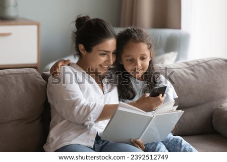 Indian mother and preschooler daughter sit on sofa at home holding storybook, distracted from reading use modern smartphone, search or buy books online via web store library, on-line bookshop concept Royalty-Free Stock Photo #2392512749