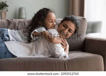 Preschooler Indian girl cuddles young beautiful mother relaxing on sofa, feel carefree, spend leisure enjoy time together and communication on weekend at modern home. Happy motherhood, cherish concept Royalty-Free Stock Photo #2392512765