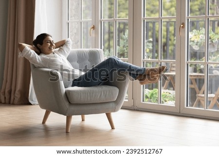 Carefree dreamy Indian woman relax on soft armchair in modern domestic room with panoramic windows, put hands behind head smiles enjoy stress-free weekend at home. Homeowner leisure, daydreams concept Royalty-Free Stock Photo #2392512767