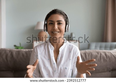 Head shot profile picture Indian woman sit on couch at home wear headphones talk to camera, webcam view. Job interview event, distance communication, business or personal virtual meeting, tech concept Royalty-Free Stock Photo #2392512777