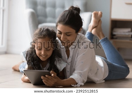 Attractive Indian preschool daughter young mother lying on warm floor in living room using digital tablet, play on-line video games, spend time on internet at home. Modern technology, leisure concept