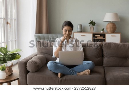 Smiling Indian woman looks at laptop screen seated on couch enjoy new video vlog, watch on-line movie spend weekend leisure at home on internet. Young generation and modern wireless tech usage concept Royalty-Free Stock Photo #2392512829