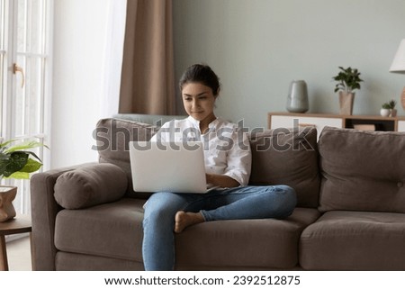 E-commerce, e-shopping, modern technology usage at home concept. Attractive Indian woman sit on sofa using laptop, do telework, looks focused, buying goods on internet, make distancing on-line order