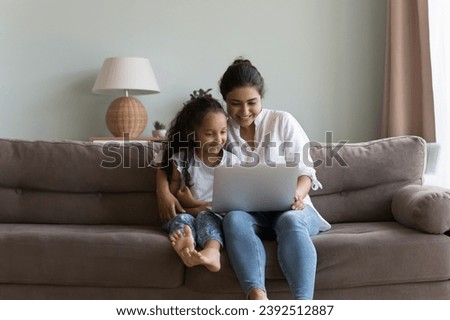 Indian woman and daughter sit on sofa with laptop, watch family movie, buying goods online spend weekend at home. Modern technology, e-commerce services, kid development use internet resources concept