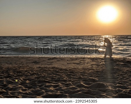 The silhouette of a little girl playing on the shore under the golden rays of the setting sun. The sea waves gently touch the shore, creating a magical atmosphere of serenity and childlike joy at dusk Royalty-Free Stock Photo #2392545305