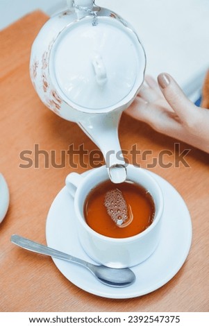Vertical image and top view of female hands of unrecognizable Caucasian woman, pouring tea in porcelain cup on table on coffee shop table