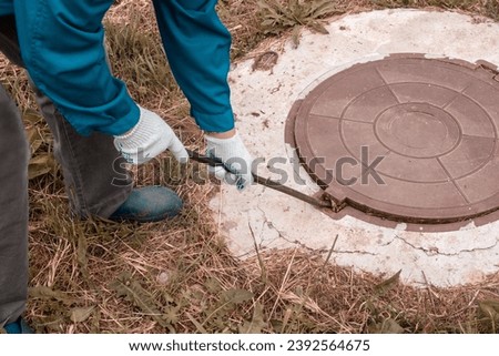 A worker opens a well hatch with a pry bar. Troubleshooting, checking septic tanks. Royalty-Free Stock Photo #2392564675