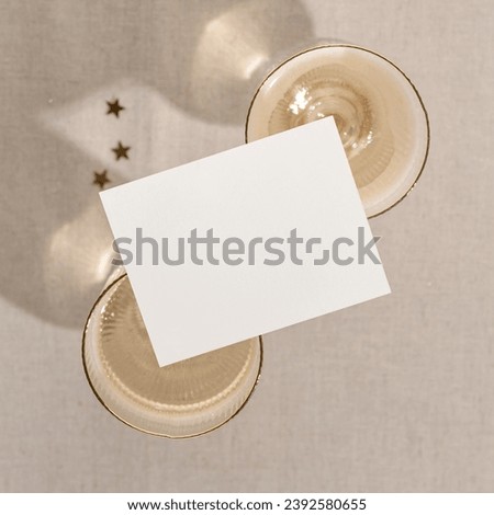 Blank paper business card mockup on wineglasses with sparkling wine, on neutral beige background with sunlight shadows. Invitation or postcard, holiday minimal branding template.