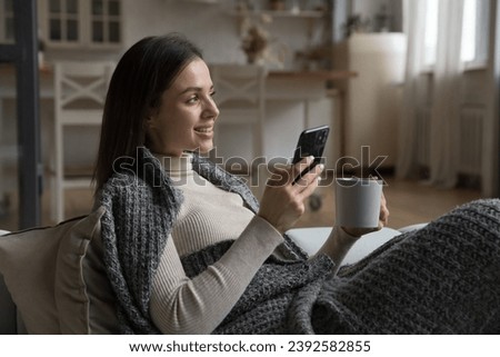 Peaceful pretty woman relaxing on couch covered with cozy plaid hold tea cup spend free time holds modern smartphone, make order, use modern tech, mobile apps for comfort life. Leisure at home concept Royalty-Free Stock Photo #2392582855
