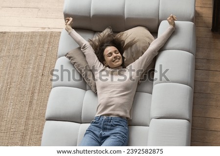Well rested woman lying on soft cushions stretching body muscles raises her arms on cozy sofa after healthy nap, top view. Stress-free leisure, modern furniture, happy homeowner relax at home concept Royalty-Free Stock Photo #2392582875