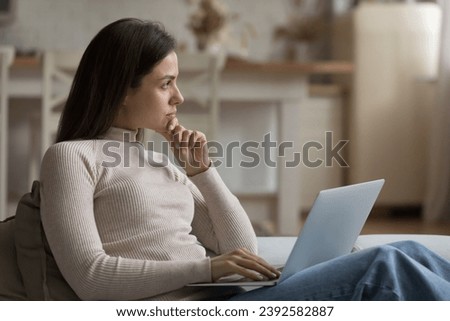 Young pensive woman working on laptop sit on sofa, looks thoughtful staring into distance deep in thoughts, thinking over task, makes telecommute job at home. Challenge, search solution, tech concept Royalty-Free Stock Photo #2392582887