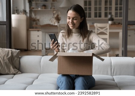 Pretty woman sit on sofa opens received courier parcel box, reviewing order in retail services using app on smart phone, check items feels happy with delivered goods. E-commerce client, tech concept Royalty-Free Stock Photo #2392582891