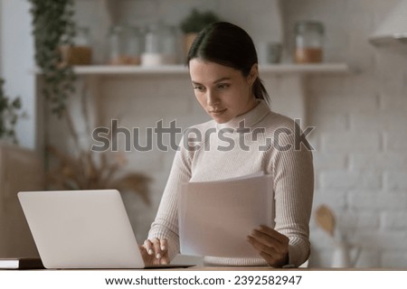 Young attractive woman do paperwork, manage utilities, paying household bills via e-bank system using laptop, looks serious sit in domestic kitchen. Telecommuting from home, modern technology concept Royalty-Free Stock Photo #2392582947