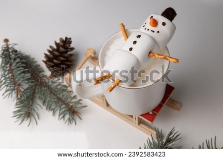 Christmas morning with a cozy cup of hot chocolate (or cappuccino) featuring cheerful snowman marshmallows . the snowman is lying in a mug. spruce branches and wooden sleigh