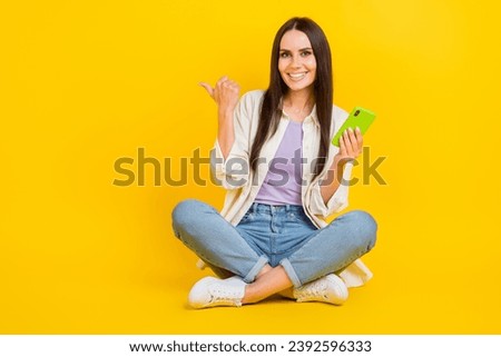 Full size photo of positive pretty girl sit floor hold smart phone indicate finger empty space ad isolated on yellow color background