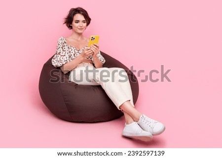 Full size photo of adorable smart girl dressed stylish blouse sit on pouf look at smartphone read email isolated on pink color background