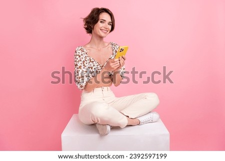 Full size photo of good mood girl dressed white trousers blouse sit on cube hold smartphone read post isolated on pink color background